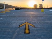 a yellow arrow painted in concrete at sunset at a parking lot with sun setting and buildings in the distance