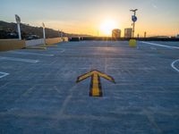 a yellow arrow painted in concrete at sunset at a parking lot with sun setting and buildings in the distance