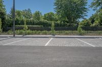 a car park with a fence, trees and paved street surface with parking spaces and white lines