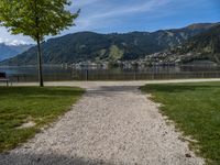 the path leads up to a park by the water, and is surrounded by grassy area with mountains in the background