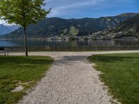 the path leads up to a park by the water, and is surrounded by grassy area with mountains in the background