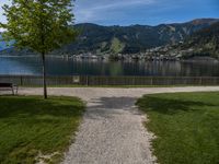 the path leads up to a park by the water, and is surrounded by grassy area with mountains in the background