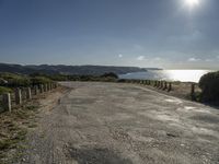 the sun shines brightly on a quiet paved area, near the ocean and a rock - fenced dirt road