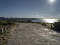 the sun shines brightly on a quiet paved area, near the ocean and a rock - fenced dirt road