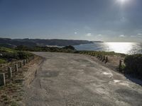 the sun shines brightly on a quiet paved area, near the ocean and a rock - fenced dirt road