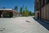 a paved concrete plaza with benches and trees in front of a building with two doors