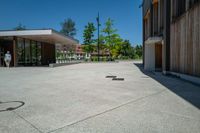 a paved concrete plaza with benches and trees in front of a building with two doors