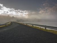 a paved road along side the water with fog covering the trees and mountains in the distance