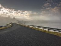 a paved road along side the water with fog covering the trees and mountains in the distance