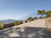 the paved road has a bench on it overlooking the sea and mountainside with trees