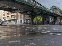 people are crossing the street under a green bridge, while rain falls on them in this photo