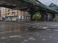 people are crossing the street under a green bridge, while rain falls on them in this photo