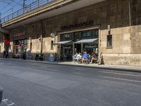 a line of people are waiting outside of a restaurant under a bridge near the city street