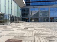 two people walking through an empty courtyard with many glass buildings in the background from the perspective of a parked motorcycle