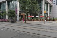 people walking on a sidewalk in front of a building next to trees and other outdoor furniture