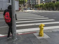 a person with a backpack standing at the corner of an intersection crossing the street near a yellow fire hydrant