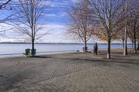 a person walking a dog next to a bench near the water with trees in the background