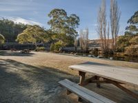 picnic tables sit in the middle of a gravel path, in a park setting and beside a river