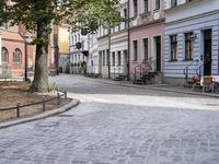 a cobblestone road leads through the middle of town where several people are eating outside a restaurant