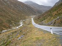 a winding country road through mountains and valleys below snow - capped peaks, a single passenger car is on the opposite side, and one person is on the right