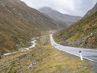 a winding country road through mountains and valleys below snow - capped peaks, a single passenger car is on the opposite side, and one person is on the right