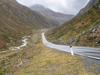 a winding country road through mountains and valleys below snow - capped peaks, a single passenger car is on the opposite side, and one person is on the right