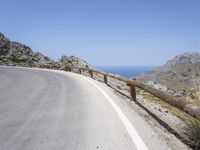 there is a wooden fence on this paved road next to the ocean and mountains on a beautiful day