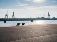 the view of a harbor with the ocean and cargo cranes in the background, in front of a couple of benches sitting on a cement area at the edge