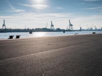 the view of a harbor with the ocean and cargo cranes in the background, in front of a couple of benches sitting on a cement area at the edge