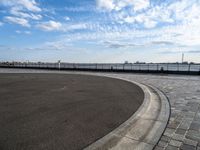 a brick walkway runs around a concrete park area next to a lake in the distance