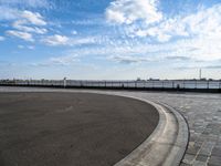 a brick walkway runs around a concrete park area next to a lake in the distance