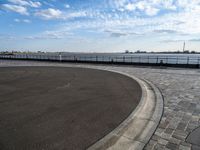 a brick walkway runs around a concrete park area next to a lake in the distance