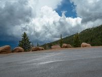 there is a black sign that is on a road with mountains in the background, on top of a hill near trees