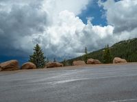 there is a black sign that is on a road with mountains in the background, on top of a hill near trees