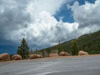 there is a black sign that is on a road with mountains in the background, on top of a hill near trees