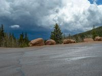 there is a black sign that is on a road with mountains in the background, on top of a hill near trees