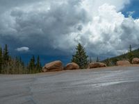 there is a black sign that is on a road with mountains in the background, on top of a hill near trees