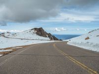 an empty road with snow and mountains in the back ground under cloudy skies on a sunny day