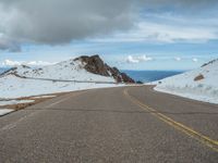 an empty road with snow and mountains in the back ground under cloudy skies on a sunny day