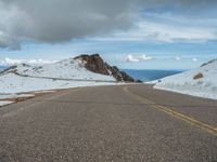 an empty road with snow and mountains in the back ground under cloudy skies on a sunny day