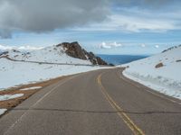 an empty road with snow and mountains in the back ground under cloudy skies on a sunny day