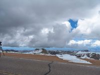 an empty road with snow and mountains in the back ground under cloudy skies on a sunny day