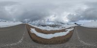 a snow covered road and snow capped mountains under a cloudy sky, taken in an upside down lens