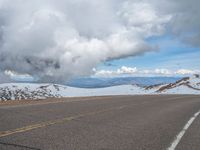 an empty road with snow and mountains in the back ground under cloudy skies on a sunny day