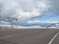 an empty road with snow and mountains in the back ground under cloudy skies on a sunny day