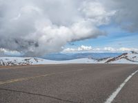 an empty road with snow and mountains in the back ground under cloudy skies on a sunny day