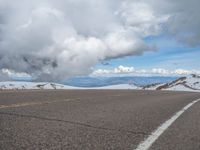 an empty road with snow and mountains in the back ground under cloudy skies on a sunny day