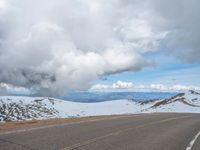 an empty road with snow and mountains in the back ground under cloudy skies on a sunny day