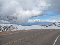 an empty road with snow and mountains in the back ground under cloudy skies on a sunny day