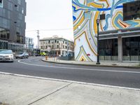 two cars driving on a street by a tall building that has an abstract mural painted on the side of it